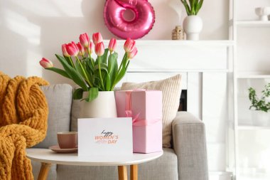 Vase with tulips, cup and greeting card for Women's Day on table in living room