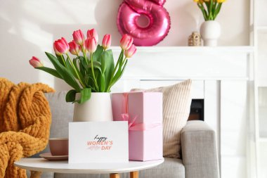 Vase with tulips, cup and greeting card for Women's Day on table in living room