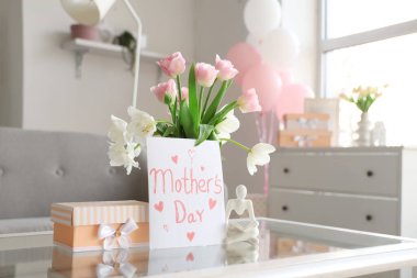 Anneler günü kartı. Oturma odasındaki masada çiçekler ve hediye kutusu var.