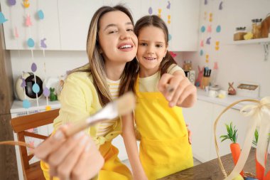 Mutlu anne ve sevimli küçük kızı neşeli mutfakta boyalı Paskalya yumurtaları için fırçalarla