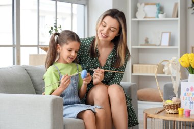 Mutlu anne ve küçük tatlı kızı ellerinde fırçalarla şenlikli oturma odasındaki kanepeye Paskalya yumurtası boyuyorlar.