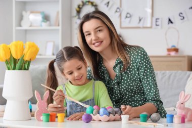 Mutlu anne, küçük tatlı kızıyla birlikte şenlikli oturma odasındaki masada paskalya yumurtası boyuyor.