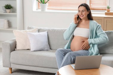 Evdeki koltukta cep telefonuyla konuşan hamile bir kadın.