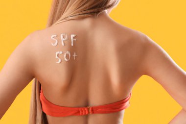 Sarı arka planda güneş kreminden yapılmış SPF 50 yazılı mayolu genç bir kadın.