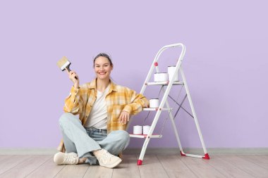 Leylak duvarının yanındaki merdiven üzerinde fırça ve boya kutuları olan genç bir kadın.