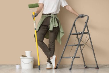 Bej duvarın yanında merdiveni ve boyası olan genç bir kadın.