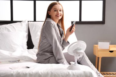 Yatak odasında cep telefonu ve elektrikli vantilatörü olan genç bir kadın.