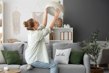 Kanepedeki genç kadın evdeki lambaya ampul takıyor.