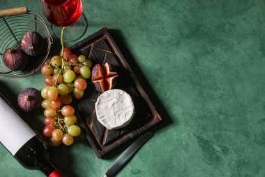 Olgun üzümlü ahşap tahta, yeşil arka planda Camembert peyniri, incir ve şarap şişesi.