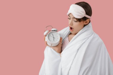 Pembe arka planda battaniyeli ve çalar saati olan uyku maskeli güzel bir kadın.