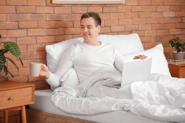 Evde yatağında çay ve dizüstü bilgisayarı olan genç bir adam.