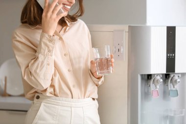 Soğutucudan su içen ve mutfakta cep telefonuyla konuşan genç bir kadın.