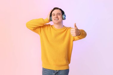 Kulaklıklı yakışıklı, genç ve mutlu bir adam. Renkli arka planda baş parmak hareketi gösteriyor.