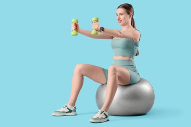 Dambılı genç bir bayan mavi arka planda fitbol antrenmanı yapıyor.