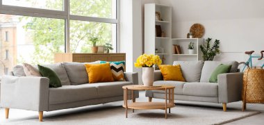 Aydınlık oturma odasının içinde gri kanepeler ve ahşap sehpanın üzerinde sarı narsisli çiçekler.