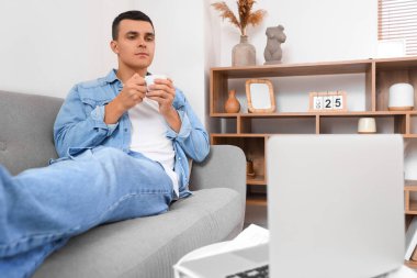 Genç adam kanepede dizüstü bilgisayarla kahve masasında yatarken espresso içiyor.