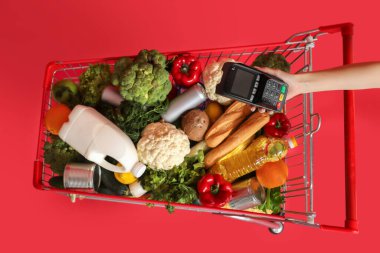 Kırmızı arka planda ödeme terminali olan yiyecek ve kadın eli dolu bir alışveriş arabası.