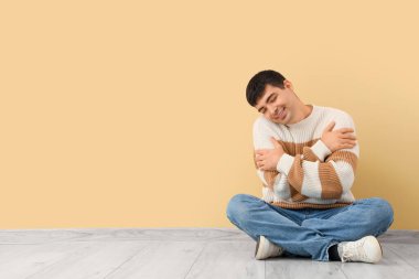 Genç adam bej duvarın yanında kendine sarılıyor.