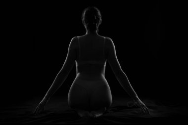 Karanlık arka planda oturan, iç çamaşırlı, çekici genç bir kadın silueti.