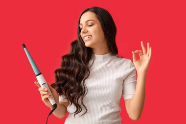 Beautiful young woman with curling iron showing OK on red background