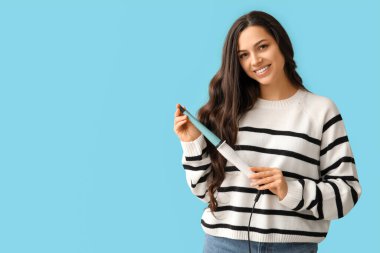 Beautiful young woman with curling iron on blue background