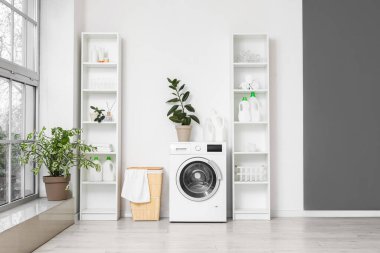 Beyaz duvarın yanında sepetli ve raf üniteli modern çamaşır makinesi. Ev çamaşır odasının içi.