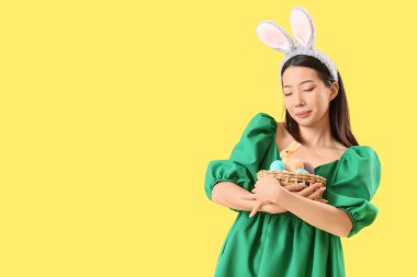Sarı arka planda Paskalya yumurtası sepeti olan tavşan kulaklı genç Asyalı kadın.