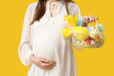 Elinde Paskalya hediyesi yumurta ve sarı arka planda oyuncak tavşan olan hamile bir kadın.