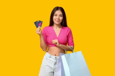 Şık kot şortlu, şık Asyalı bir kadın alışveriş torbaları ve sarı arka planda kredi kartları var.