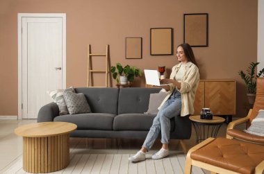 Bej oturma odasında dizüstü bilgisayarla kanepede oturan genç ve güzel bir kadın.