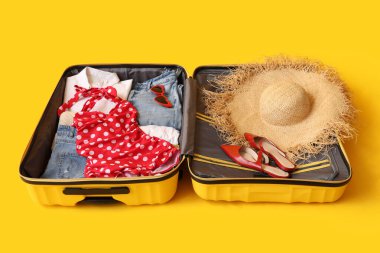 Sarı arka planda kadın kıyafetleri, şapka ve ayakkabıları olan bavulu aç. Seyahat kavramı