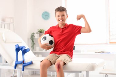 Yaralı diziyle ve futbol topuyla gülümseyen küçük çocuk klinikteki koltukta kaslarını gösteriyor.