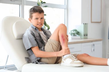 Klinikteki kanepede oturan yaralı bacaklı küçük çocuk.