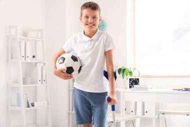 Klinikte koltuk değneği ve futbol topuyla gülümseyen küçük çocuk.