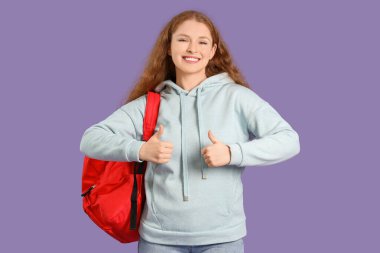 Sırt çantalı mutlu kız öğrenci, leylak arkaplanda baş parmak gösteriyor.