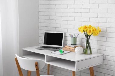 İşyerindeki beyaz tuğla duvarın yanında narsisli çiçekler ve dizüstü bilgisayarla aydınlık ofis.