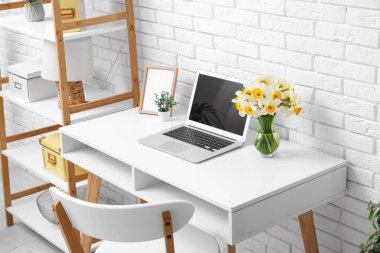 Ofisteki beyaz tuğla duvarın yanında bir buket narsisli çiçek ve dizüstü bilgisayarla raf ve işyeri.