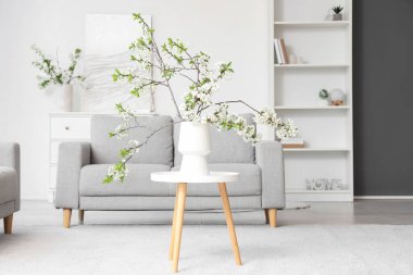 Beyaz oturma odasındaki gri kanepenin yanındaki sehpanın üzerinde çiçek açan bir vazo.