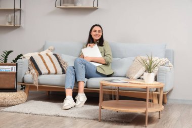 Dizüstü bilgisayarı olan genç bir kadın evdeki ışık duvarının yanındaki gri koltukta oturuyor.