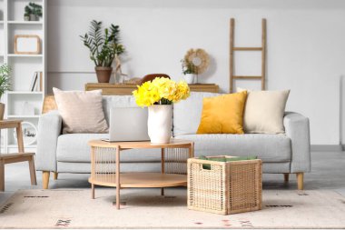 Gri kanepe, dizüstü bilgisayar ve ahşap sehpada sarı narsisli çiçeklerle oturma odasının içinde.