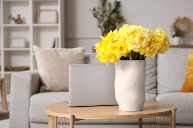 Gri kanepe, dizüstü bilgisayar ve ahşap sehpada sarı narsisli çiçeklerle oturma odasının içinde.