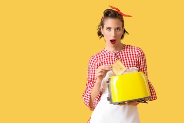 Güzel, genç, şok olmuş ev hanımı tost makinesi ve ekmek dilimiyle sarı arka planda.