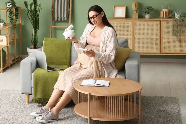 Domuz kumbarası ve hesap makinesi olan hamile bir kadın oturma odasında kanepede oturuyor.