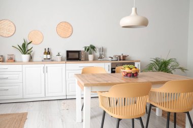 Modern mutfağın içinde beyaz tezgahlar, yemek masası, meyveler, sandalyeler ve lamba var.