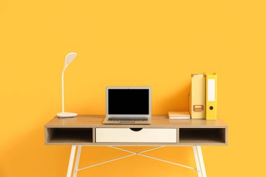 Ofiste sarı duvarın yanında dizüstü bilgisayarı ve lambası olan işyeri