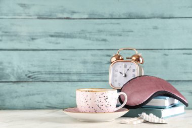 Bir fincan çay, uyku maskesi, kitaplar, haplar ve çalar saat beyaz masada.