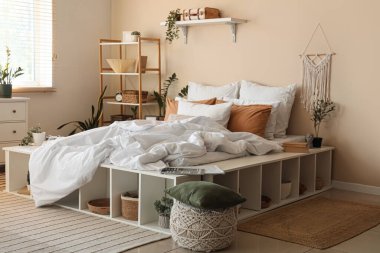 Büyük konforlu yatak, ev bitkileri, puf ve raf üniteleri yatak odasının içinde.