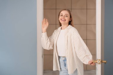 Güzel genç bir kadın açık kapıda duruyor ve el sallıyor.