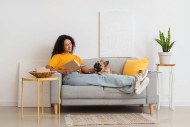 Güzel bir Afro-Amerikalı kadın oturma odasındaki kanepede tatlı bir Fransız bulldoguyla kitap okuyor.