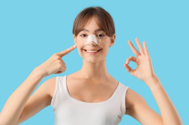 Güzel, genç, mutlu bir kadın sargılı burnu işaret ediyor ve mavi arka planda iyi bir hareket gösteriyor. Estetik ameliyat konsepti